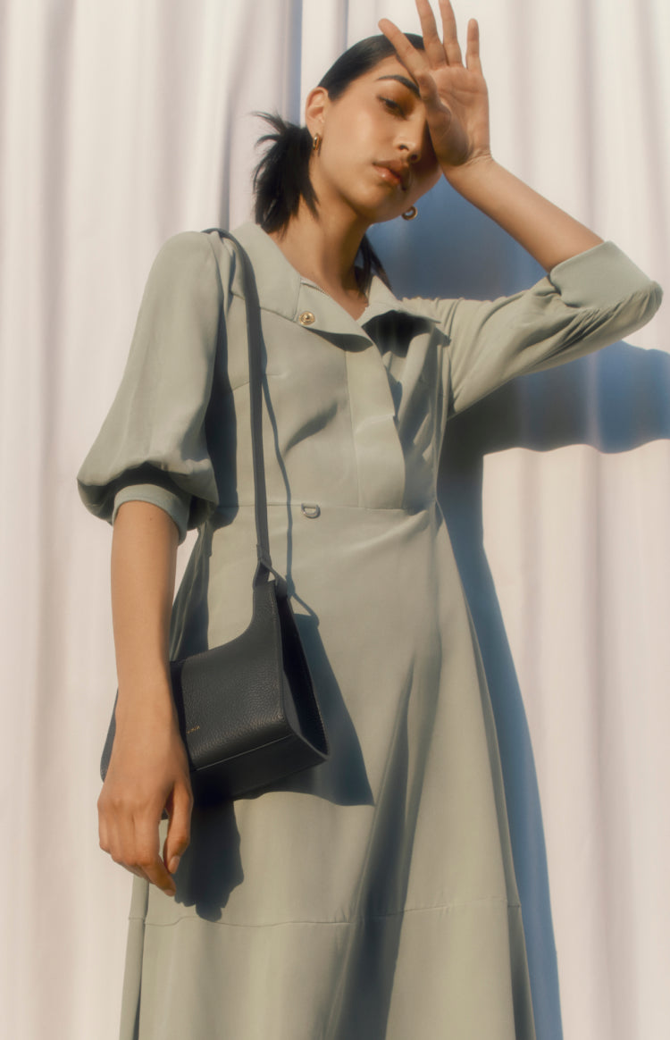 Woman in a dress with a bag posing with hand on forehead.