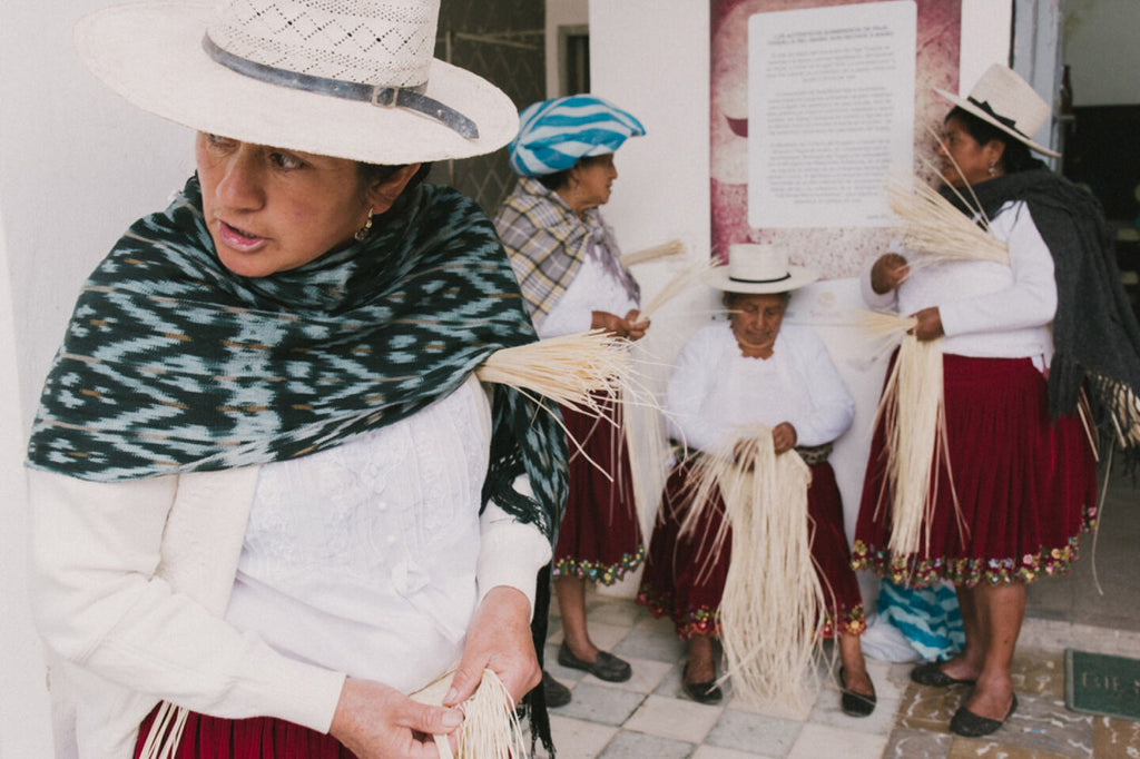 Four women, wearing hats and shawls, stand near a building working with fibers.