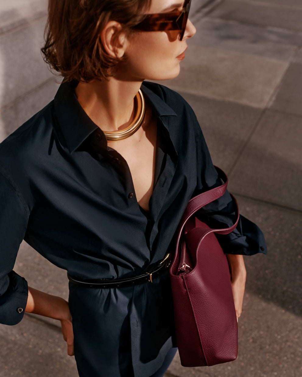 Person wearing a collared shirt with a belt, sunglasses, gold necklace, and holding a tote