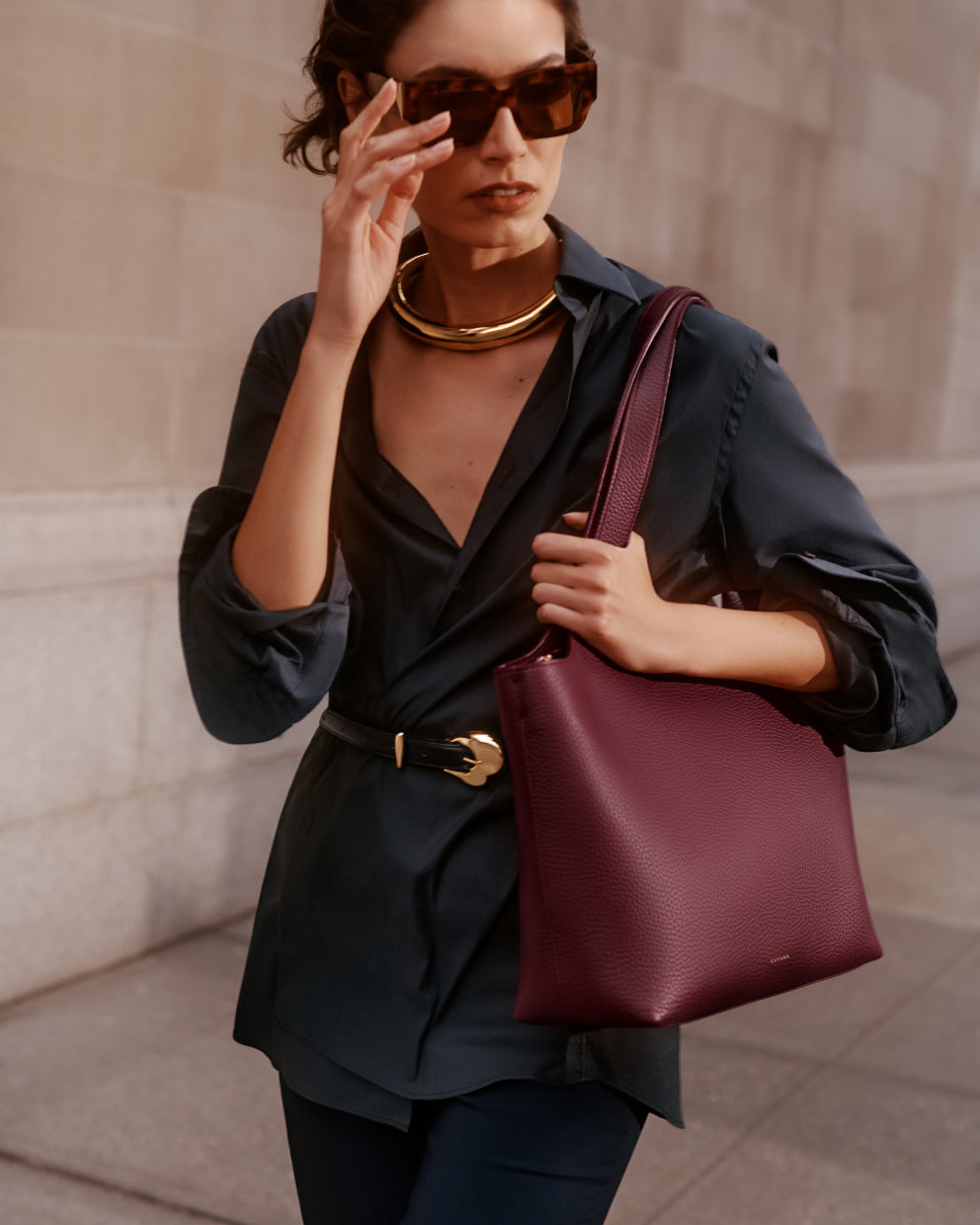 Person wearing a collared shirt with a belt, sunglasses, gold necklace, and holding a tote