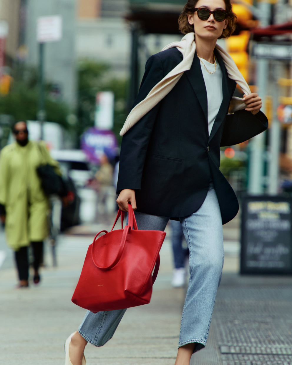 Person walking in city holding a red handbag, wearing a coat with a sweater draped over shoulders and wearing jeans