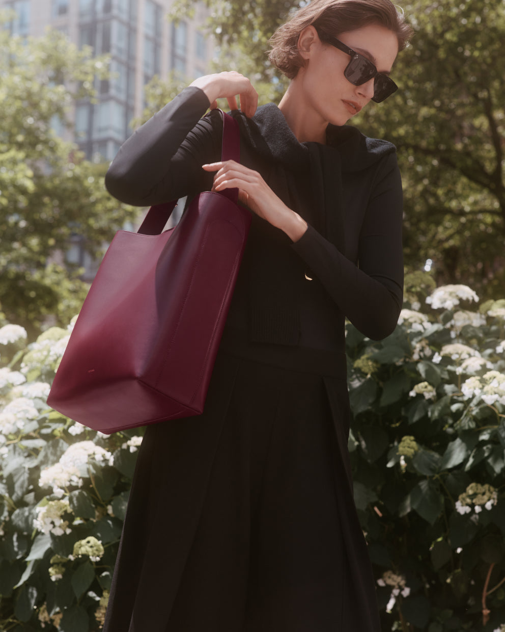 Woman in sunglasses holding a large handbag in a garden setting.