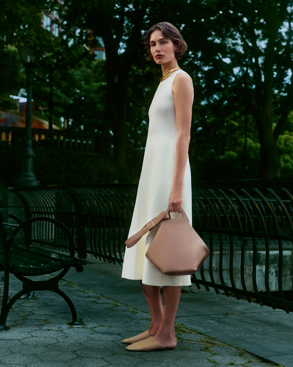 Person in sleeveless dress holding a handbag, standing near a park bench at dusk.