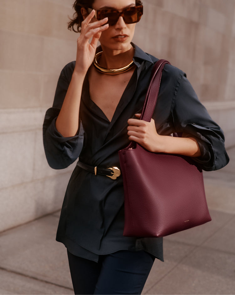Person wearing sunglasses and a necklace holding a handbag by a wall.