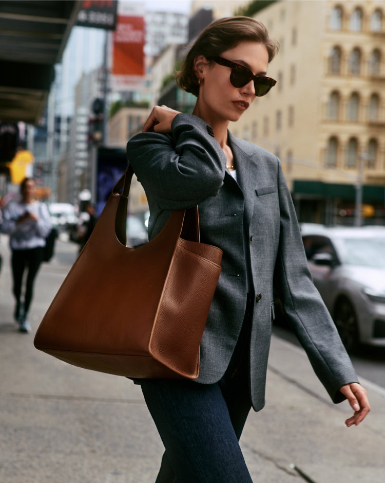 Person walking in city carrying a large tote bag wearing sunglasses and a blazer.