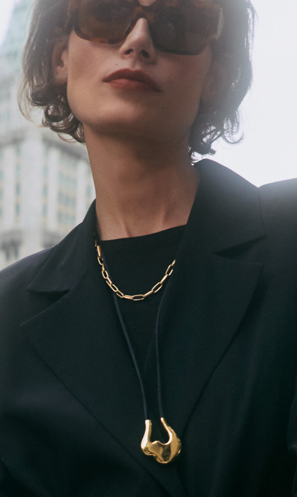 Person wearing sunglasses, blazer, gold chain necklace, abstract pendant, and dark top.