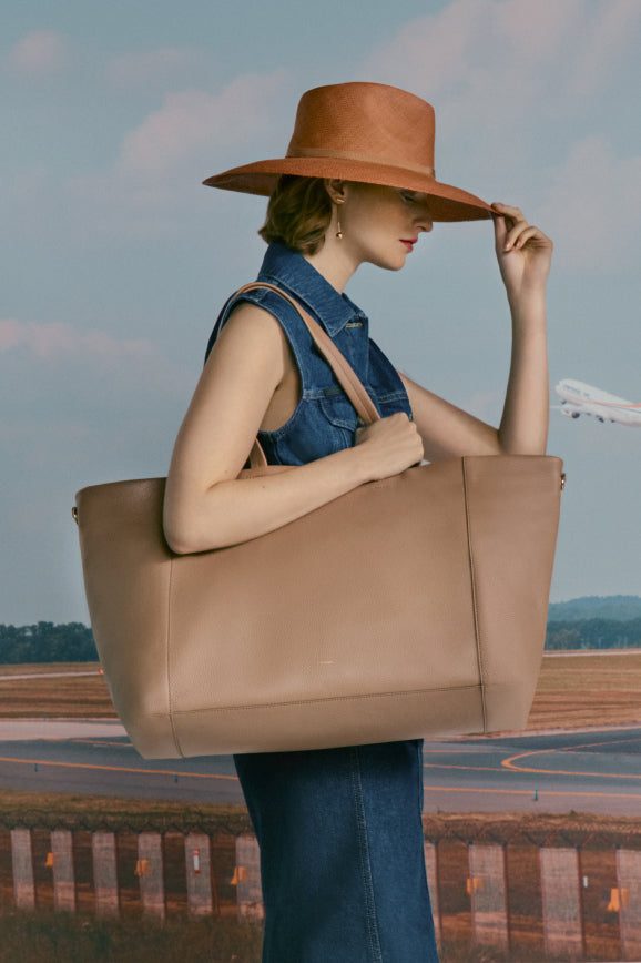Woman in a large hat holding a big bag with an airplane in the background.