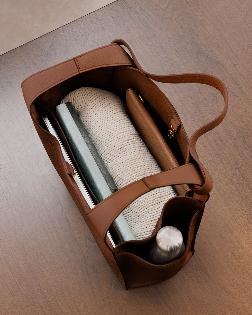 Double Loop Satchel Top view of an open handbag containing a book, a notebook, a rolled-up sweater, a wallet, and a water bottle.