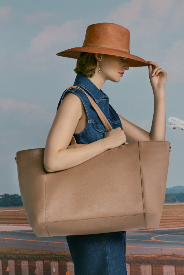 Woman holding her hat, carrying a large bag, near an airstrip.