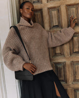 Person in a cozy sweater and skirt, holding a shoulder bag, reclining against a wooden door.