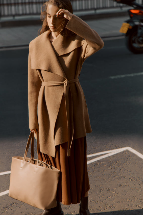 Woman in urban setting wearing a belted coat and holding a large bag.