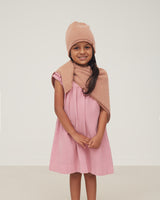 Young girl smiling, wearing a dress, beanie, and scarf, standing against a plain background.