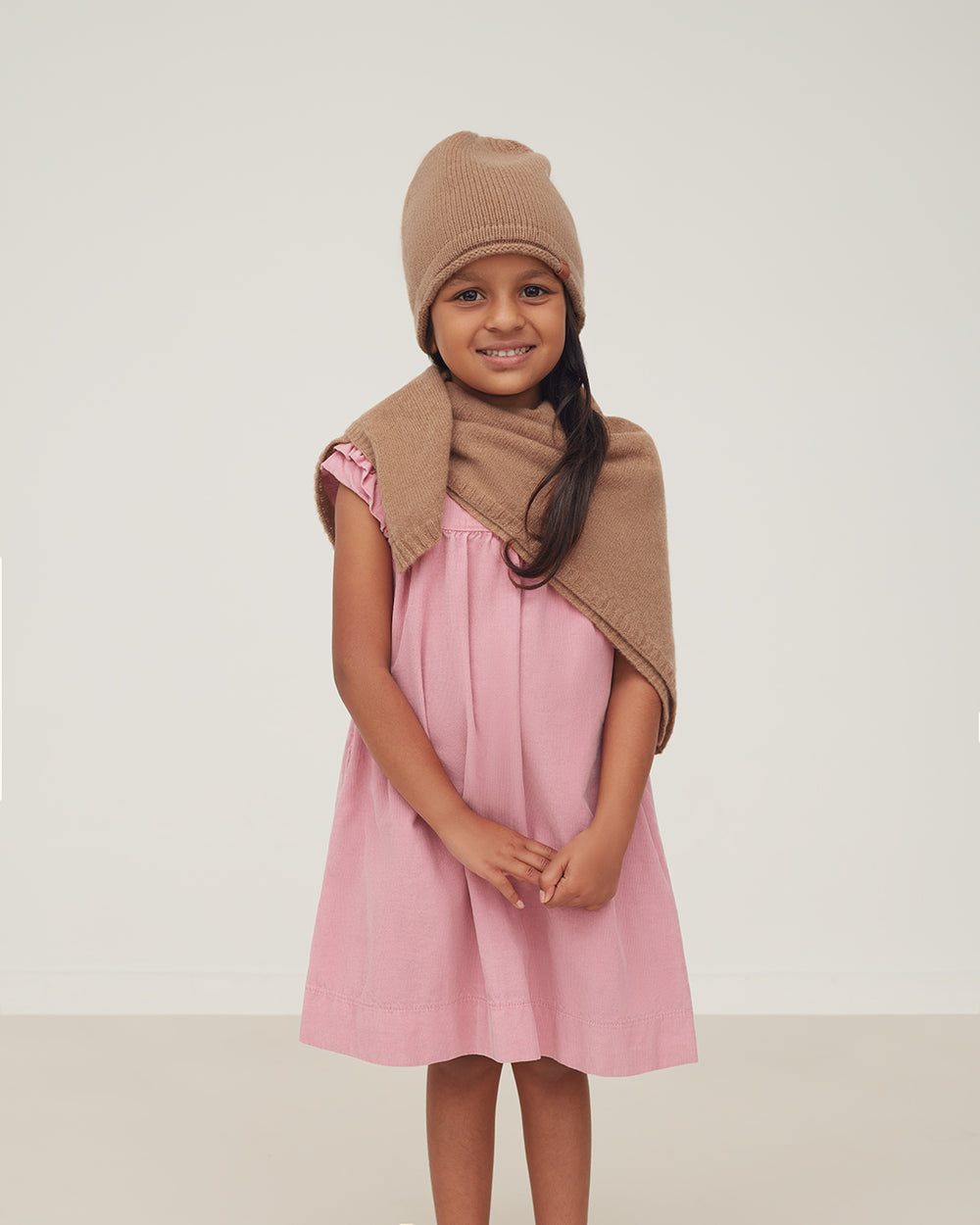Kids Single-Origin Cashmere Beanie Young girl smiling, wearing a hat and scarf, standing in a studio setting.