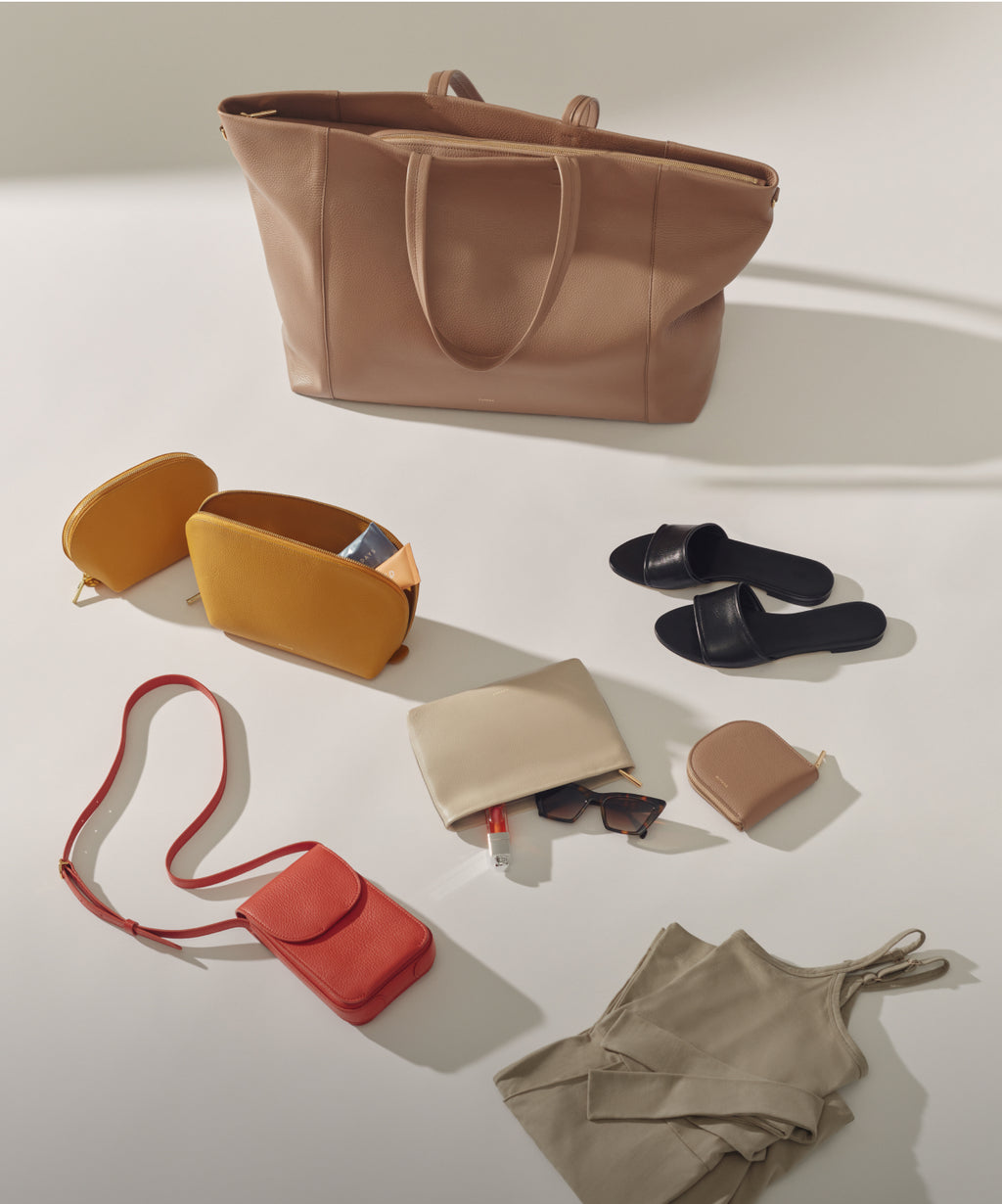 Cuyana Easy Travel Tote in color Cappuccino (top), folded clothing and leather accessories in sage, yellow, red, beige, and black. 