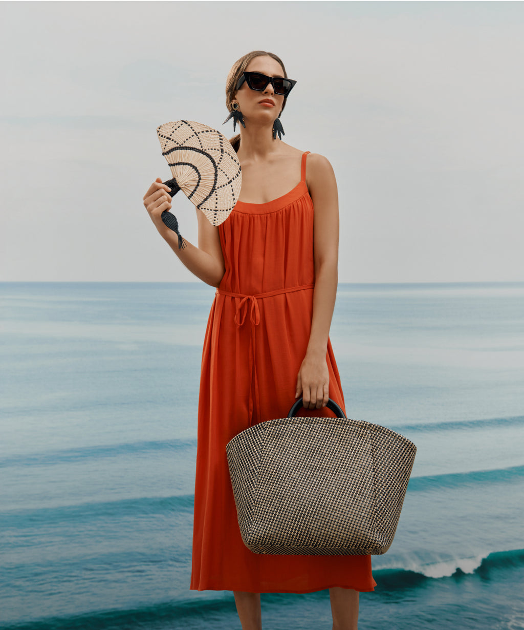 Linked Image. Model wearing orange dress, holding a straw fan with colors beige and black and large straw tote with beige and black.