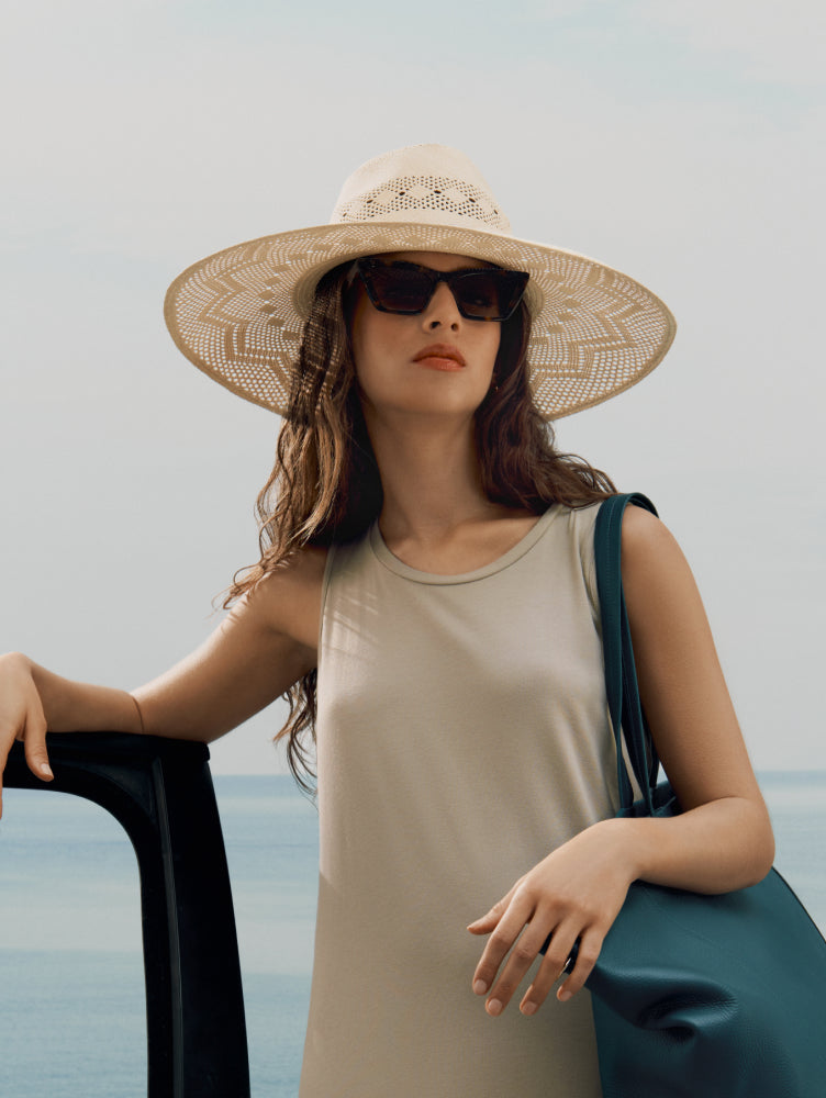 Person in sleeveless dress wearing sunhat and sunglasses, holding travel bag by car near water.