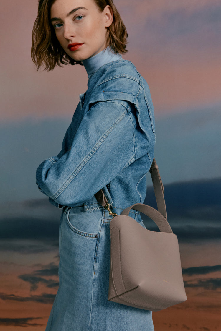 Woman in denim outfit carrying a handbag, standing against a cloudy sky.