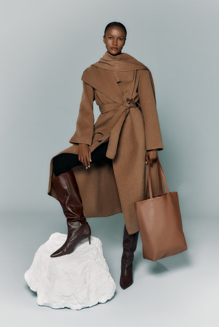 Woman standing with one foot on a rock, wearing a coat, boots, and holding a bag.