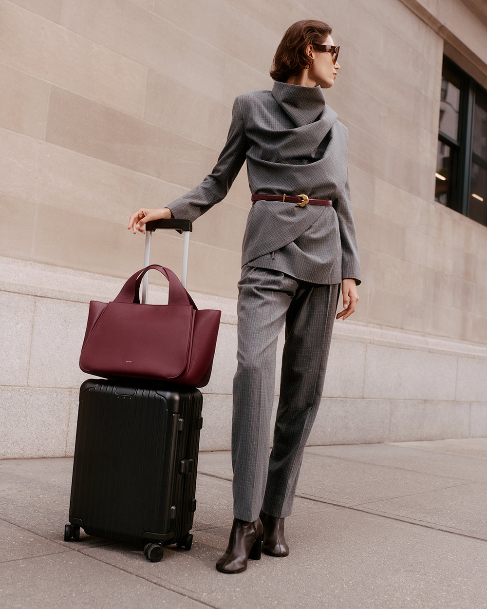 Mila Satchel Person standing with luggage and holding a large bag, wearing a belted suit and sunglasses