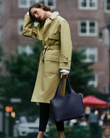 Person in a belted coat holds a large handbag in an urban setting.