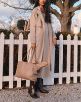 Woman in long coat holding tote bag standing by a white picket fence.