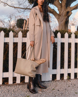 Woman in long coat holding tote bag standing by a white picket fence.