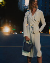 Person in a long coat holding a handbag on a city street at night.
