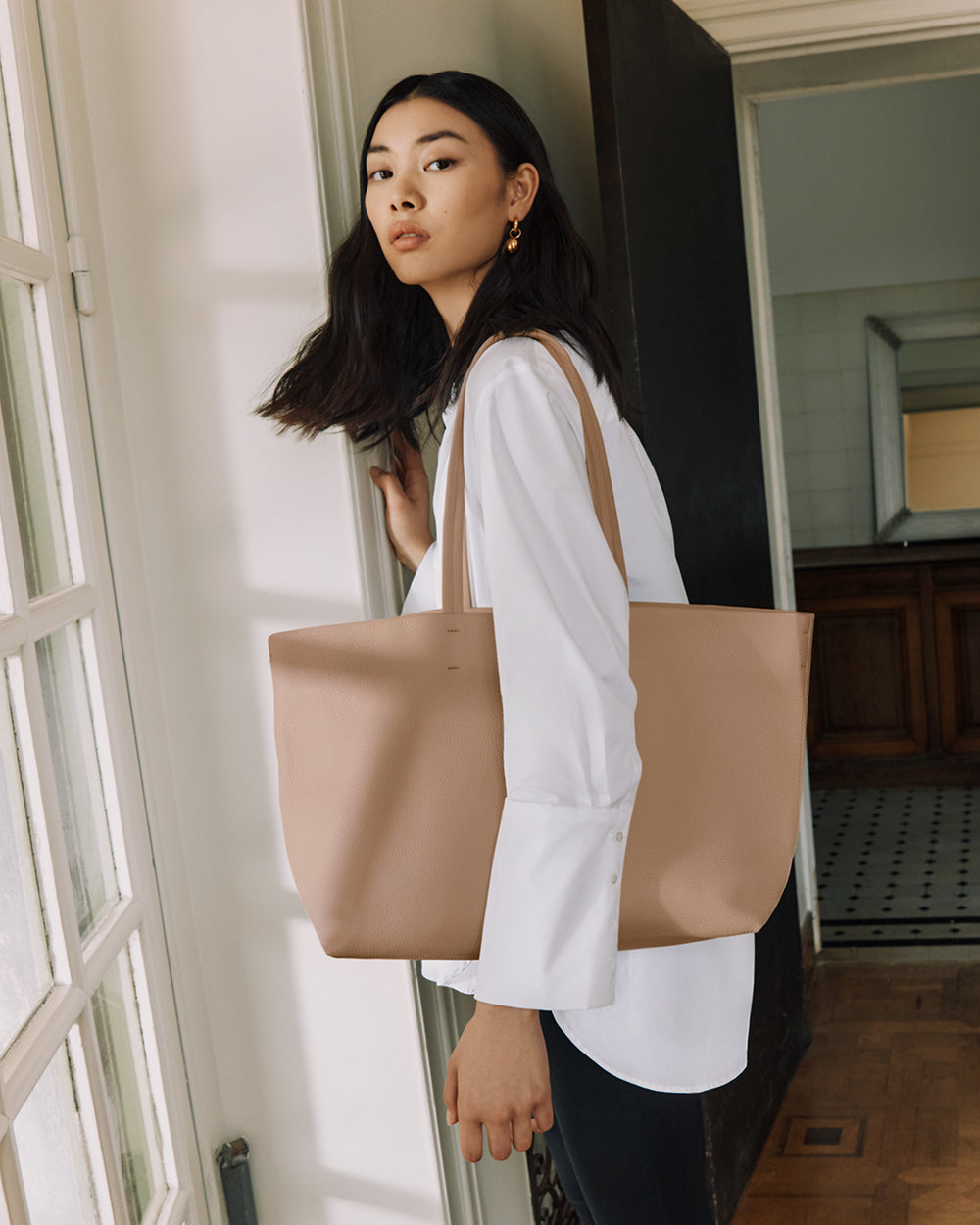 Person holding a large tote bag standing by a window indoors.