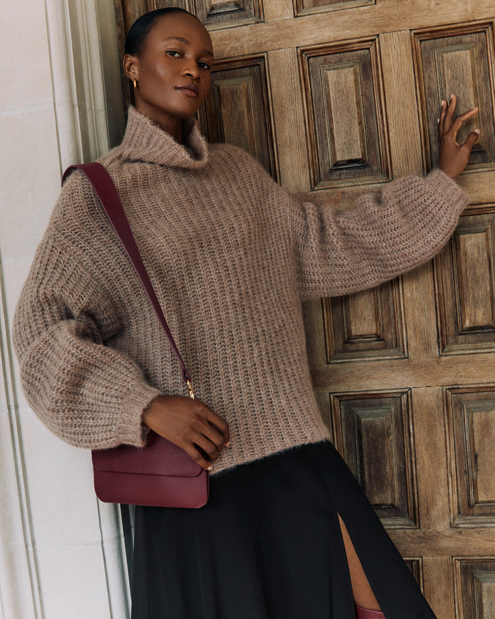 System Flap Bag Person leaning against a door wearing a sweater and skirt, with a shoulder bag