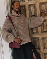 Person leaning against a door wearing a sweater and skirt, with a shoulder bag