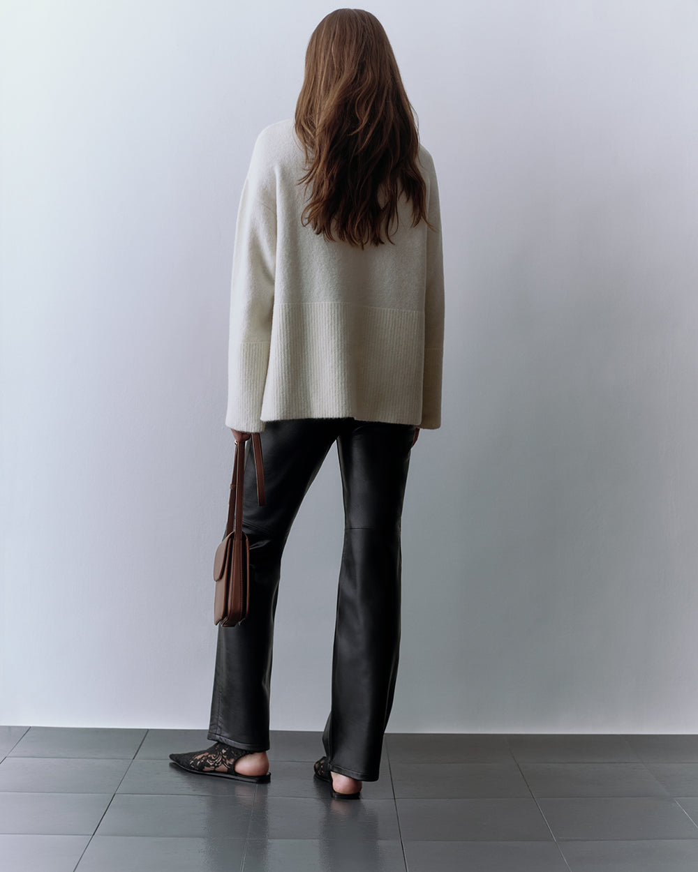 Person standing with back turned, holding a shoulder bag, wearing a sweater and pants.