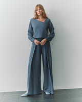 Person wearing long-sleeve top with asymmetrical neckline and wide-leg pants standing indoors.