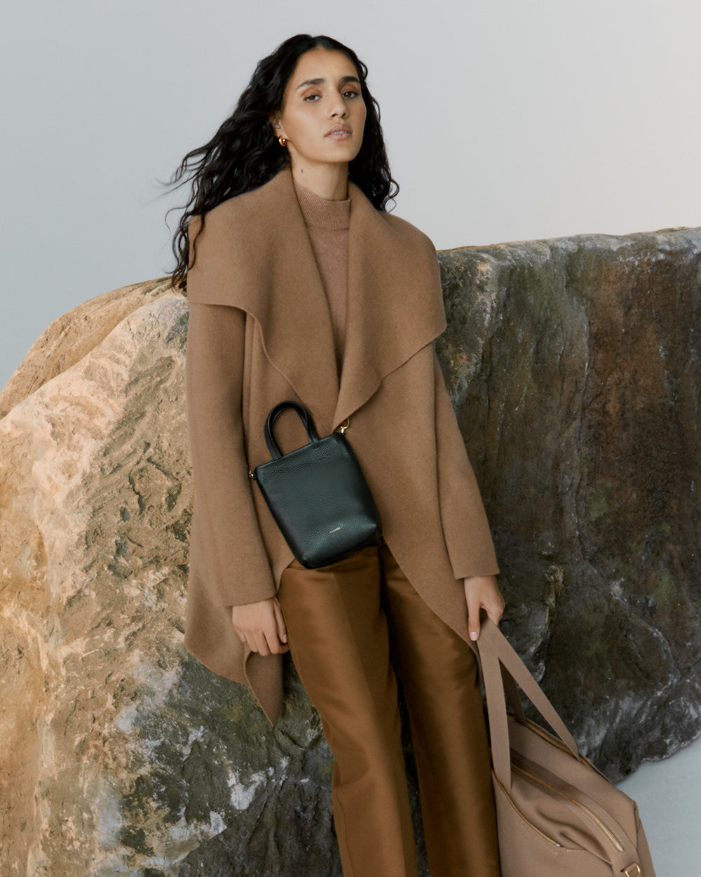 Woman standing next to a large rock, holding a bag, wearing a long coat.