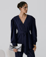 Woman standing holding a clutch, wearing a jumpsuit with a tied waist.
