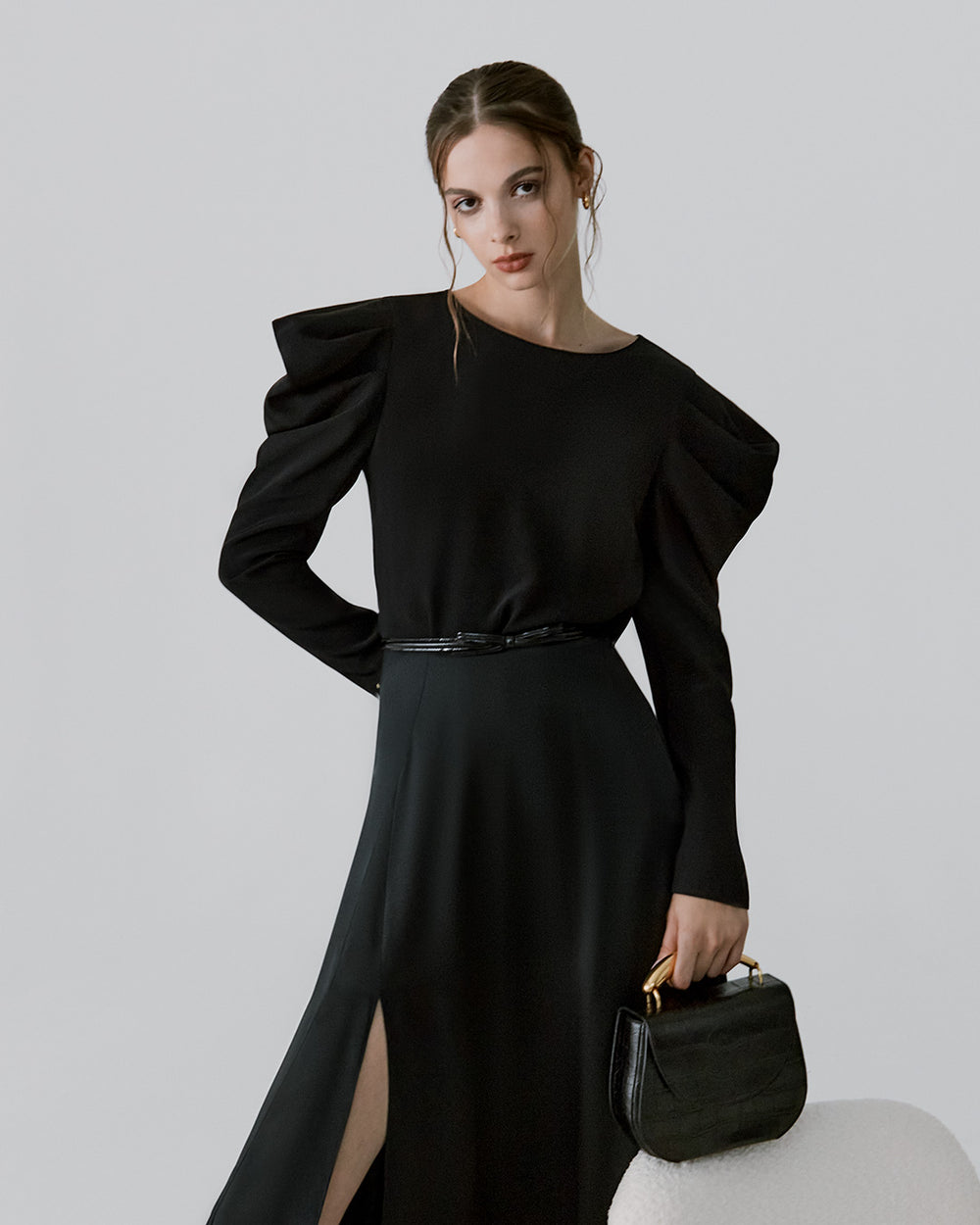Woman posing with hand on hip holding a small bag, wearing a long dress and earrings.