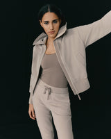 Woman in a casual outfit raising one arm, standing against a dark background.