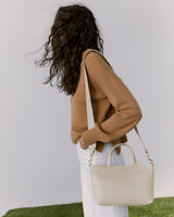 Woman standing with purse, facing away, on a grassy and plain background.