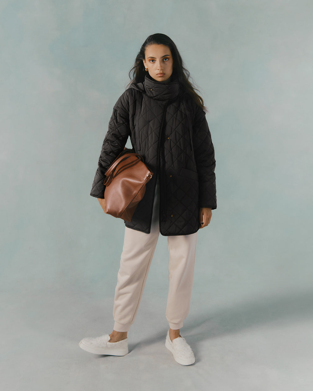 Woman standing with a coat, scarf, pants, holding a bag, and wearing sneakers.