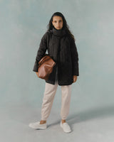 Woman standing with a coat, scarf, pants, holding a bag, and wearing sneakers.