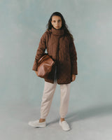 Woman standing in studio wearing a quilted jacket, holding a bag, and looking at the camera.