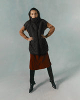 Person standing with hands on hips wearing a quilted vest, long-sleeve top, skirt, and knee-high boots.