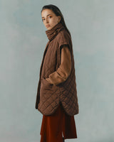 Woman in quilted jacket over dress, standing, looking at camera.