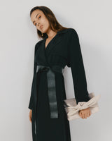 Person in a belted coat holding a clutch bag, posing against a plain background