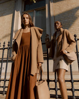 Two women posing near a building fence, one holding a bag.