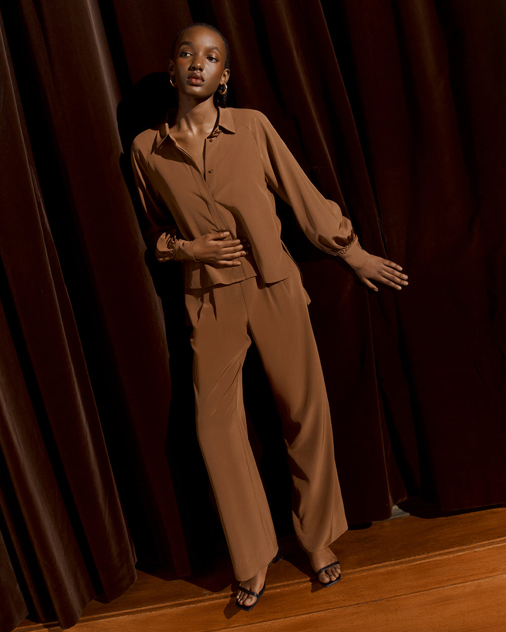 Silk Leather Cuff Shirt Woman standing in a room with curtains, wearing a buttoned shirt and pants.