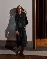 Woman standing in room with shadow on wall, wearing a dress and boots.