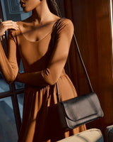 Woman in dress stands by a door with a shoulder bag.