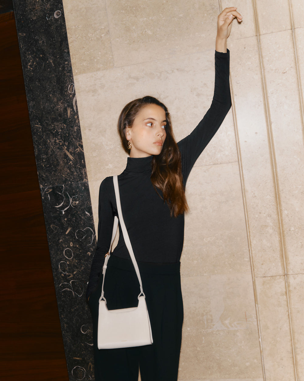 Slim Back Slit Turtleneck Tee Woman standing and reaching up against a textured wall with a bag over her shoulder.
