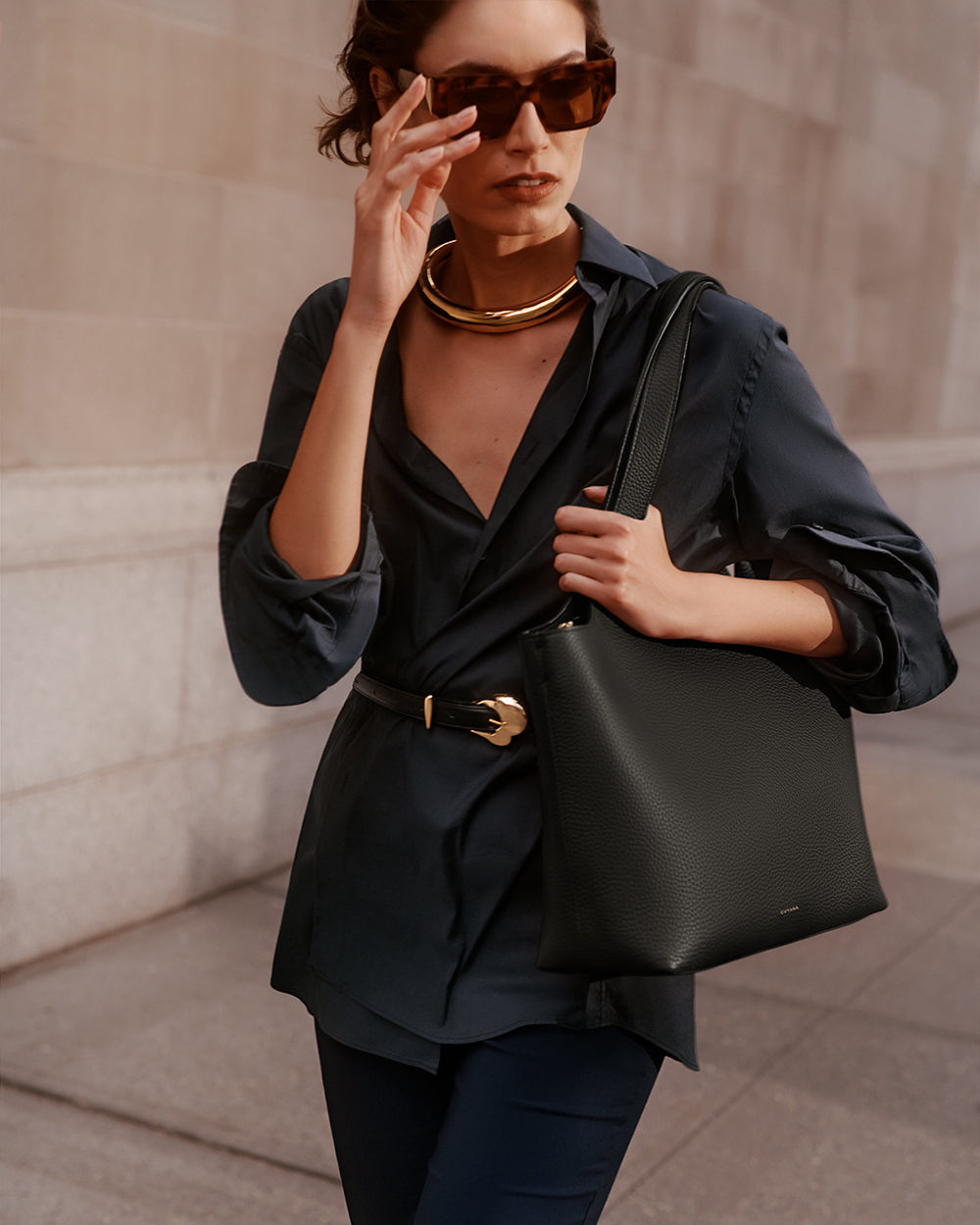 Person wearing sunglasses, holding a bag, and dressed in a blouse and pants, walking outside a building