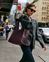 Person walking on city street carrying a large handbag, wearing sunglasses and a blazer.